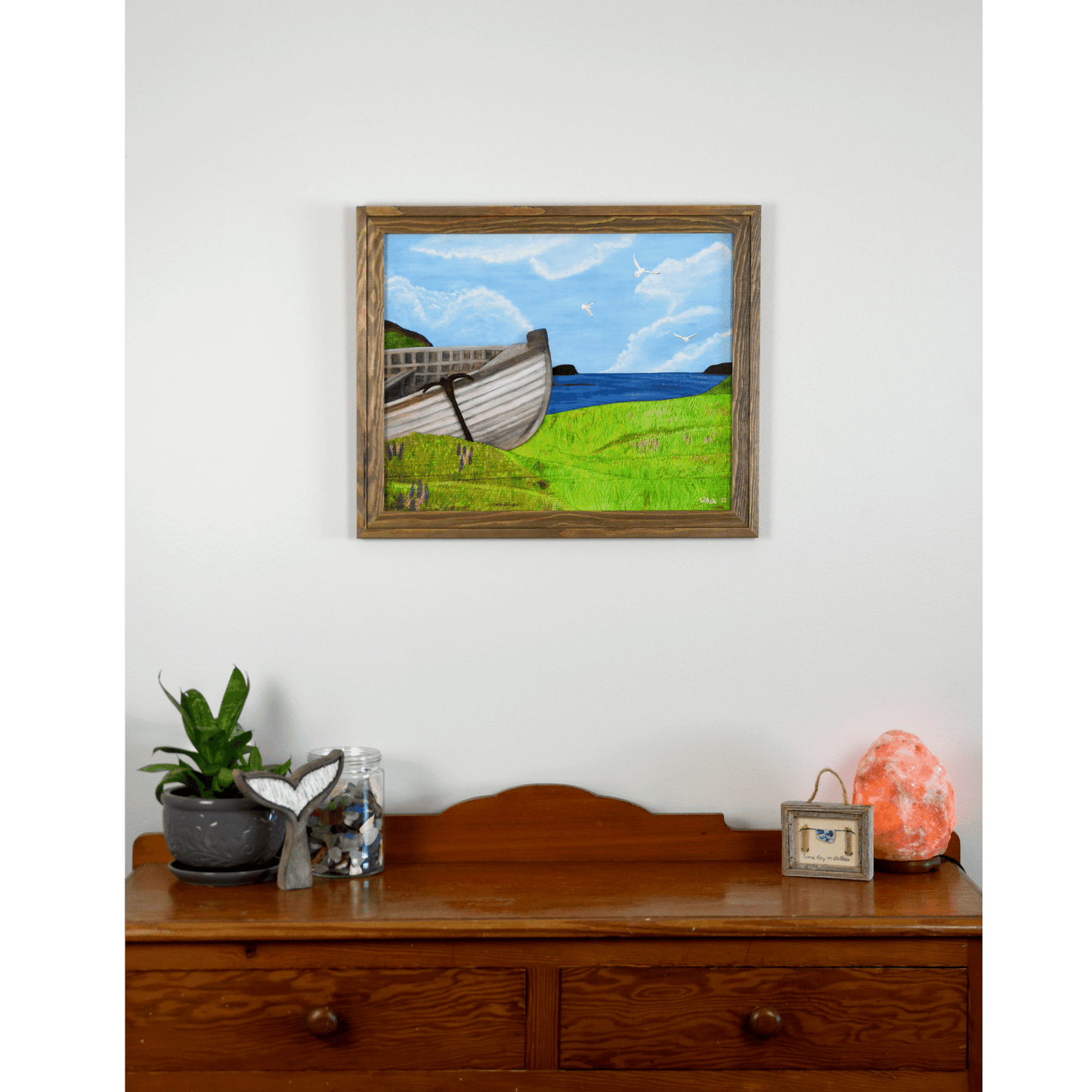 "New Beginnings" is a print featuring a wooden fishing boat that sits on a green Newfoundland shore with the ocean and seagulls in the background.