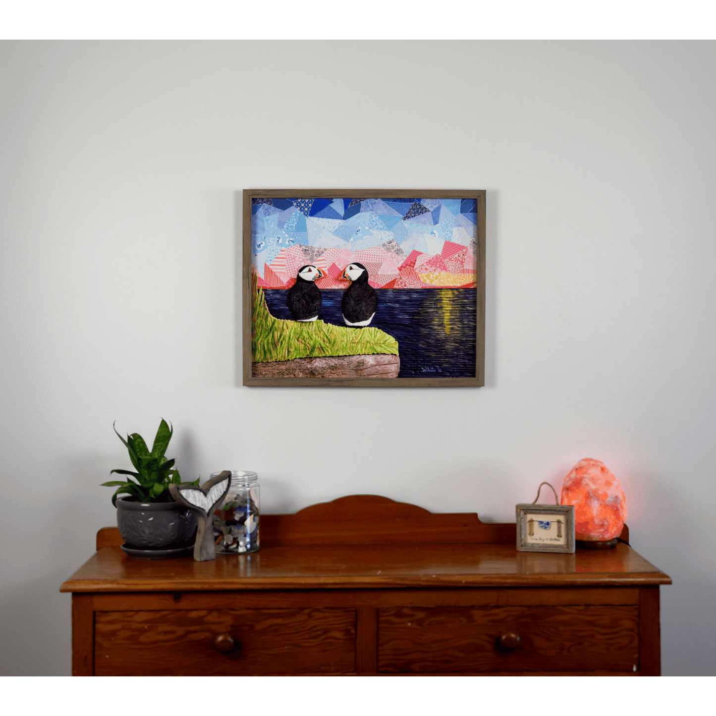 "The Perfect View" reproduction print showcases two puffins perched on a coastal rock, gazing lovingly at each other while enjoying a sunset over the ocean.