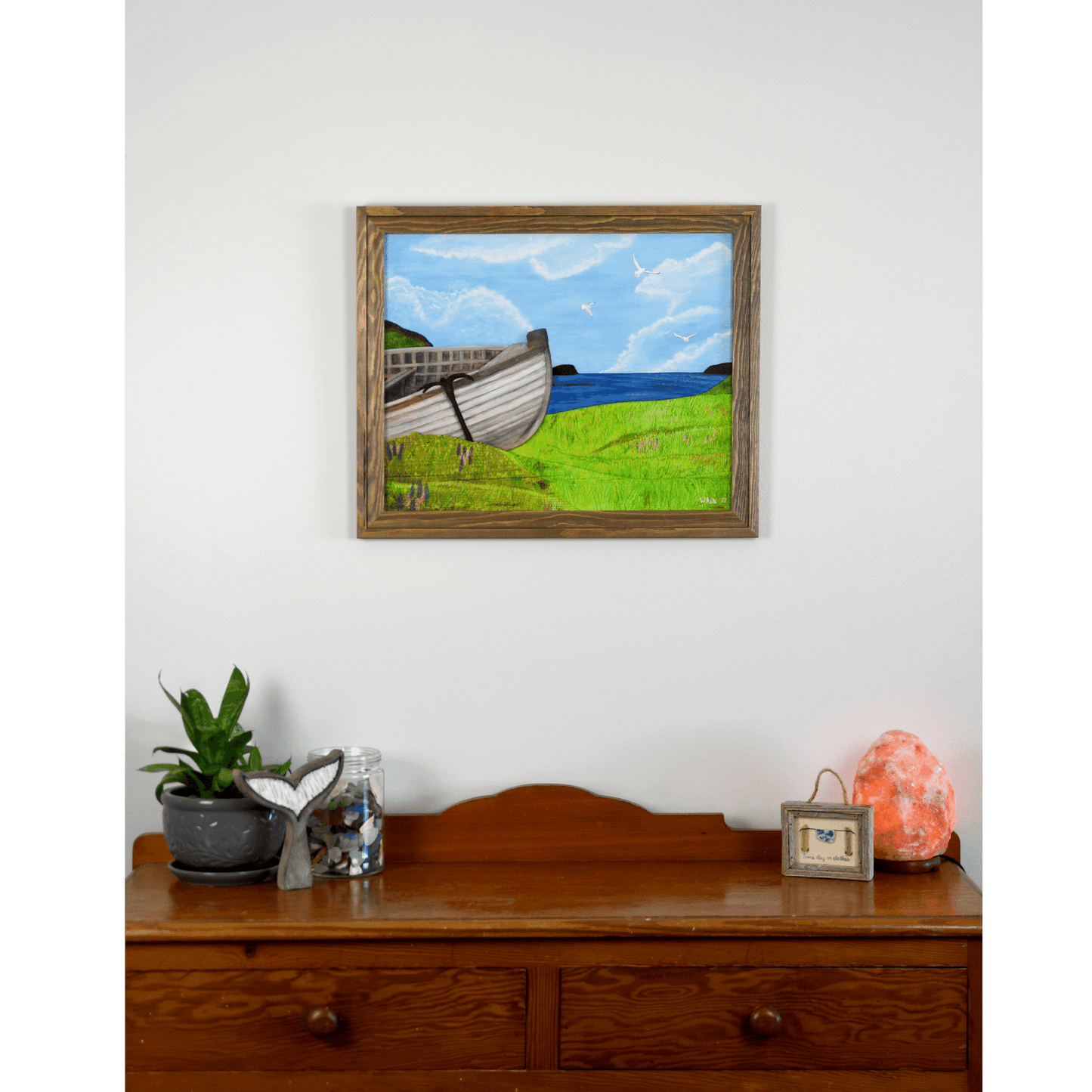 "New Beginnings" is a print featuring a wooden fishing boat that sits on a green Newfoundland shore with the ocean and seagulls in the background. 